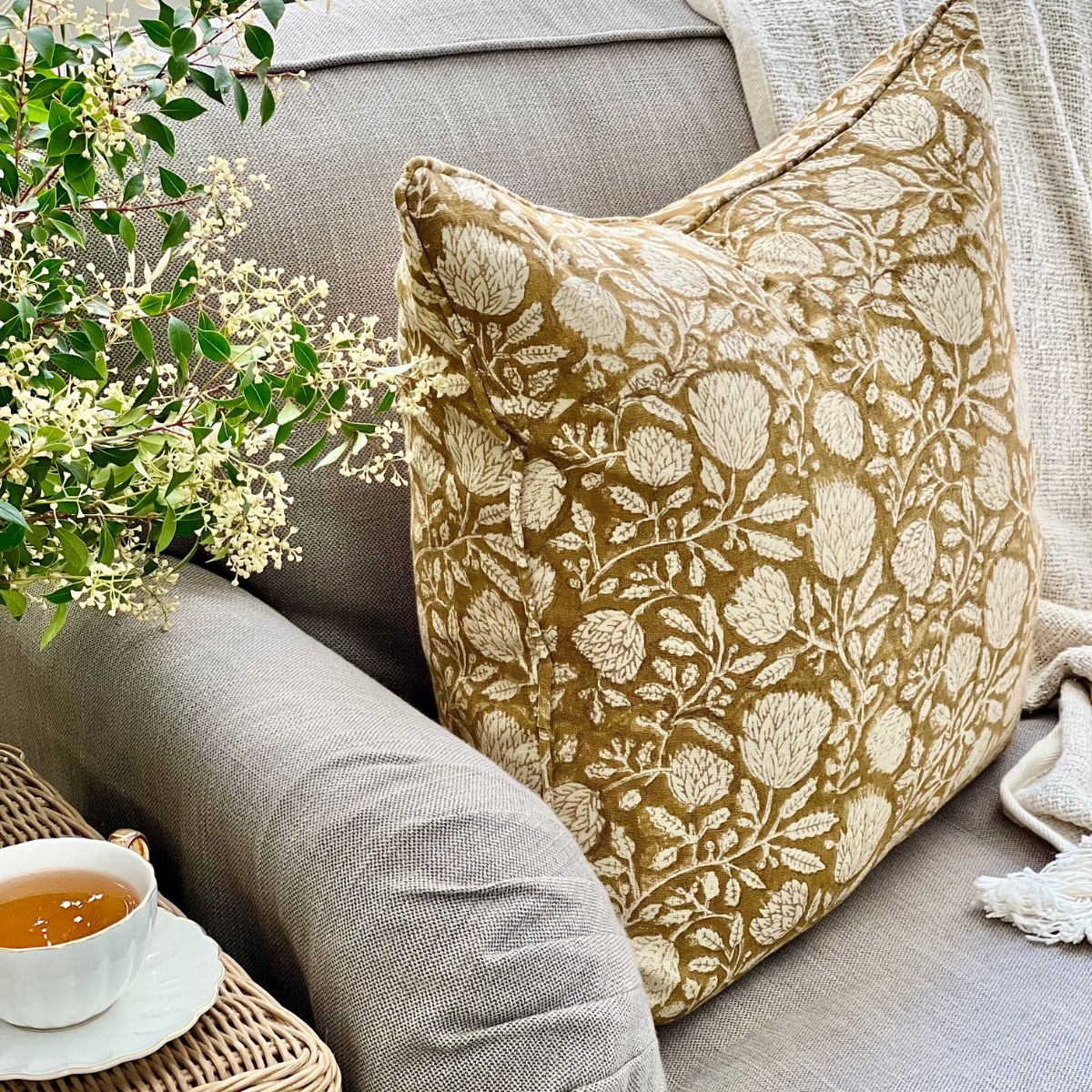 
A Golden Flora mustard monochrome floral hand block printed pure linen cushion cover with zip closure and pipe edge finish, angled on a sofa in a styled living room setting.