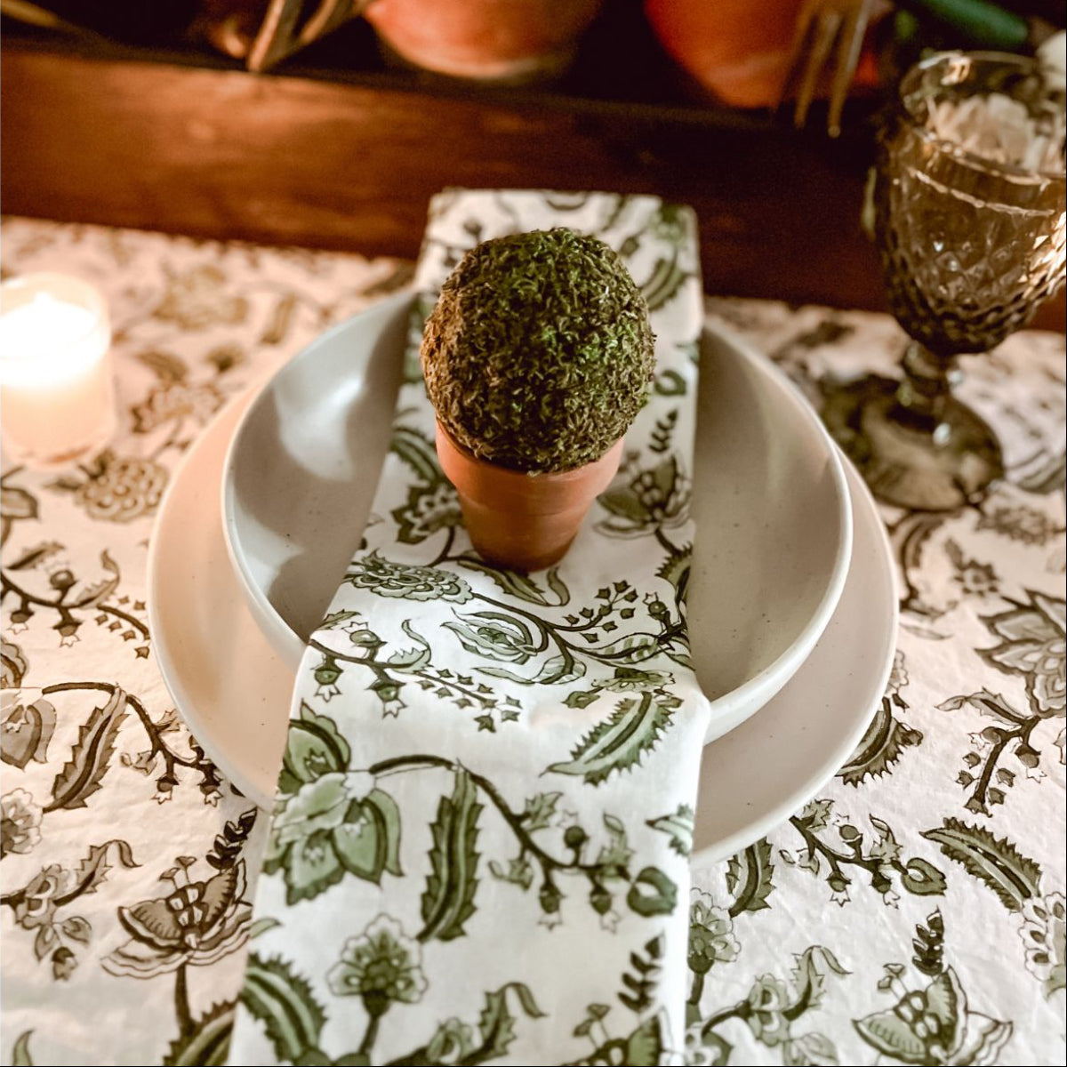 A green chintz hand block printed cotton napkin on a casual everyday dining table, styled with small potted plants for a fresh relaxed place setting.
