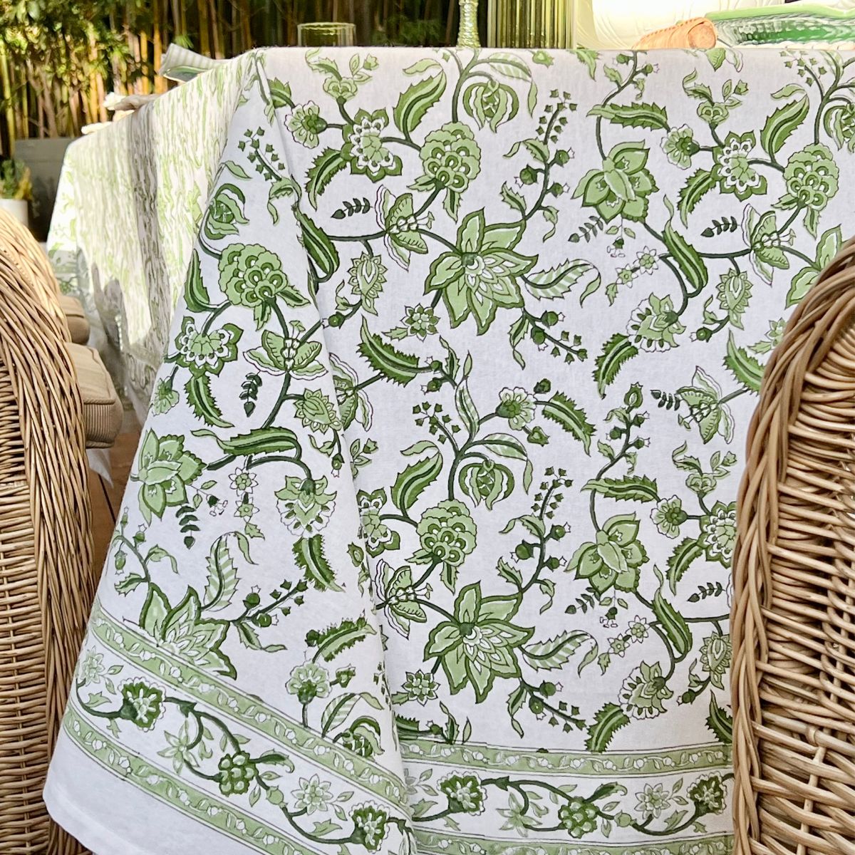 A green and white hand block printed chintz patterned square tablecloth displayed over a wicker table.