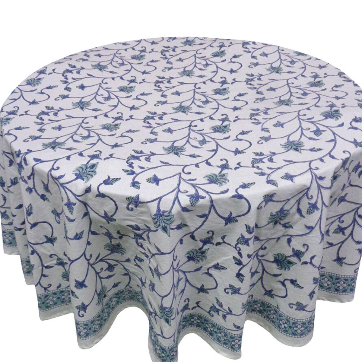 A round tablecloth with a blue floral hand block printed pattern displayed on a table accompanied by a blue and white ceramic vase and glass candle holders.