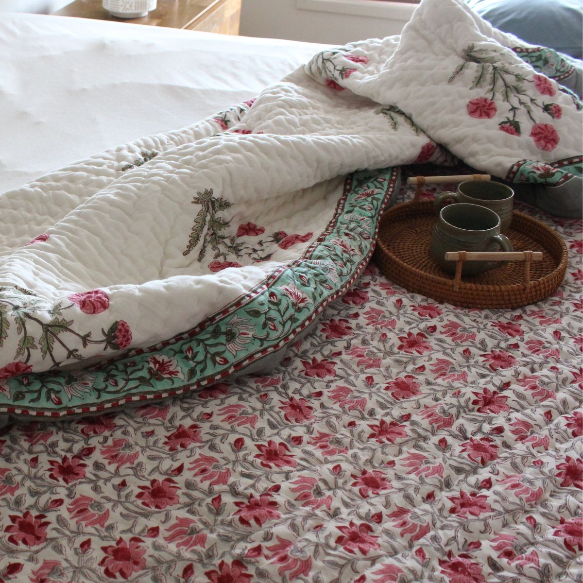 A reversible cotton quilt with pink and grey floral hand block printed patterns on a bed, with a wooden tray and cup on top of it.