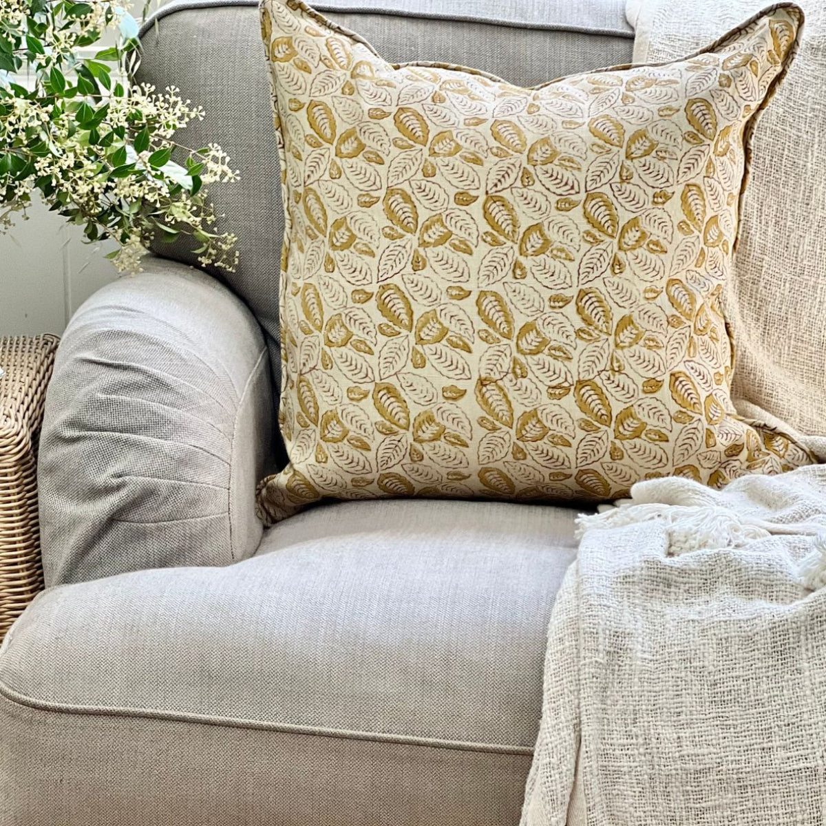 
A 50 x 50 cm Lumina Leaf mustard hand block printed pure linen cushion cover with pipe edge finish, styled upfront on a sofa with a coffee and floral vase.