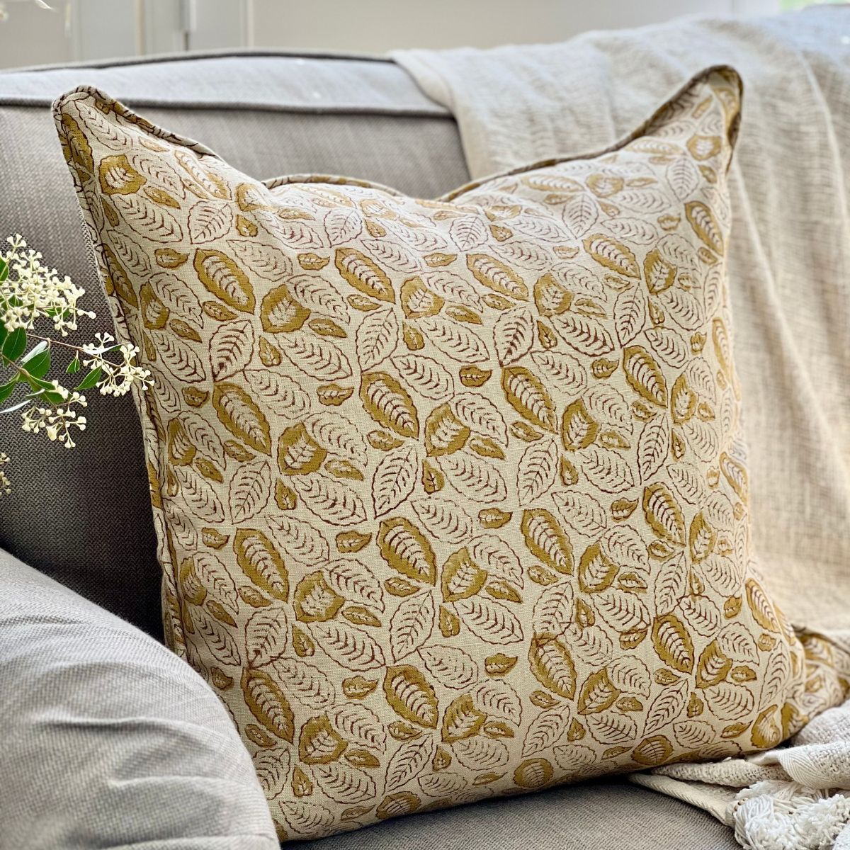 
A Lumina Leaf mustard leaf pattern hand block printed pure linen cushion cover with zip closure and pipe edge finish, angled on a sofa in a styled living room setting.