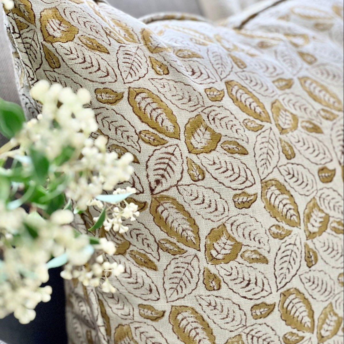 
A 50 x 50 cm Lumina Leaf mustard hand block printed pure linen cushion cover with pipe edge finish, styled upfront on a sofa with a coffee and floral vase.