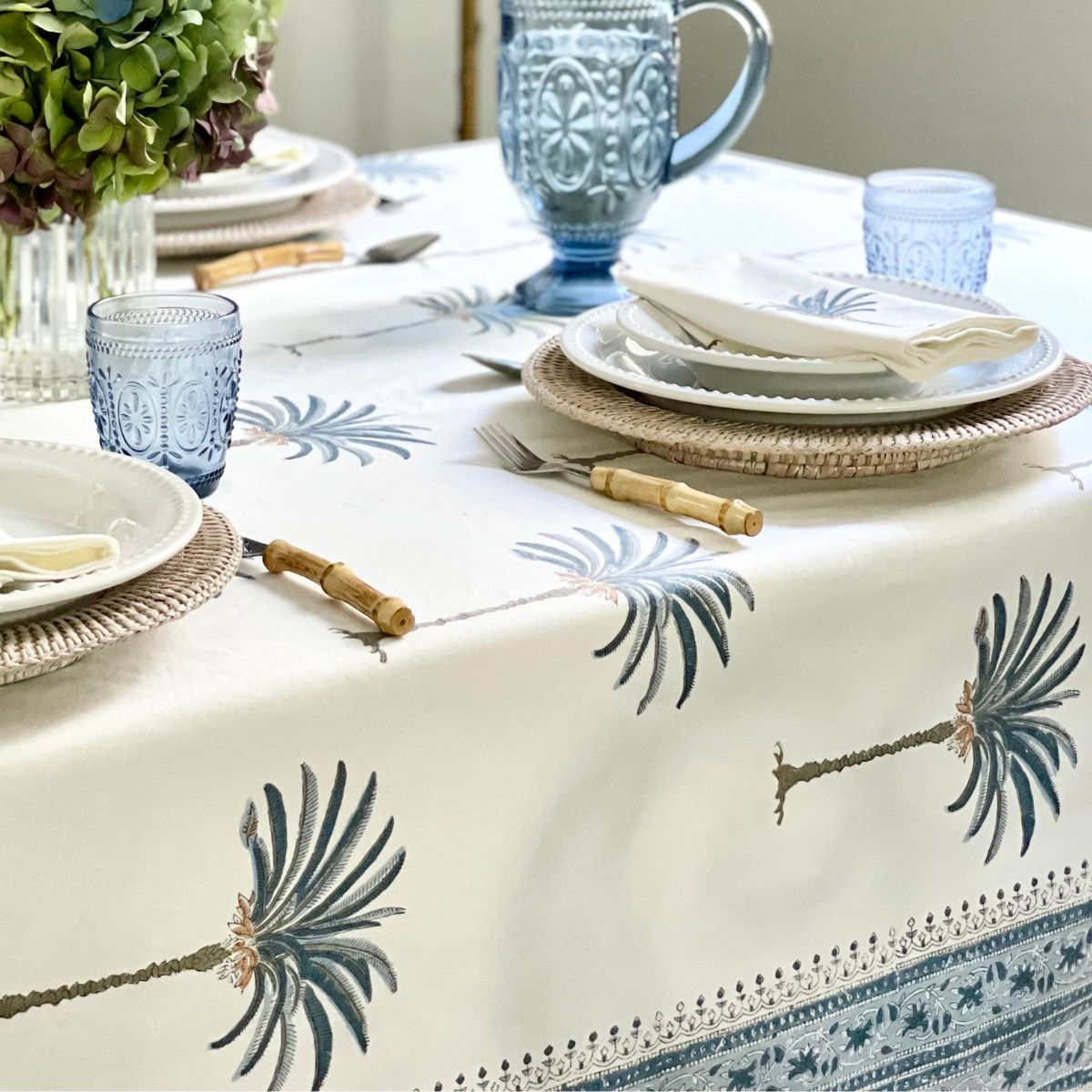 A bright casual table on a rectangular blue palm hand block printed tablecloth with an intricate border, for a fresh and elegant beach house brunch.