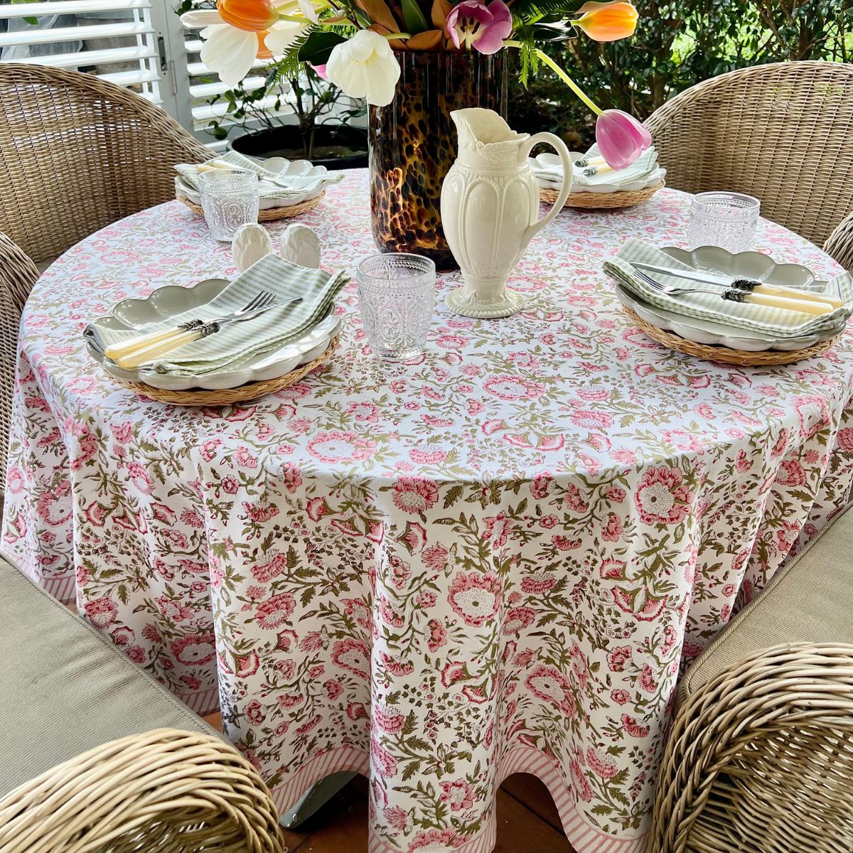 A round tablecloth with a hand block printed floral pattern in shades of pink and green, displayed on a table set with dishes and a floral arrangement.