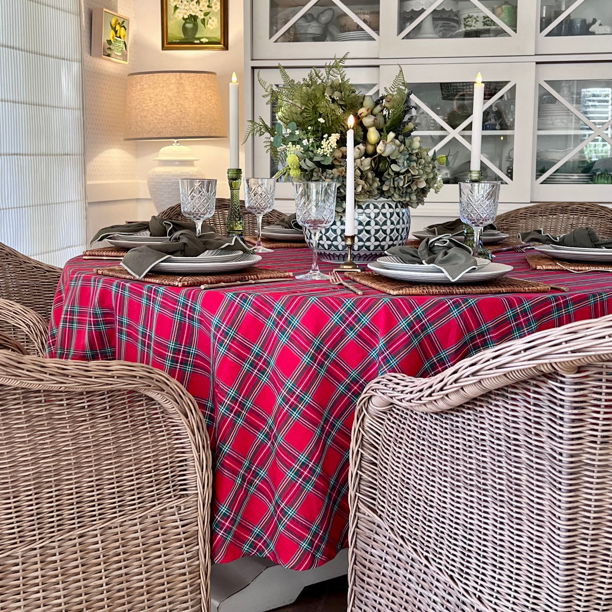 Red tartan plaid round tablecloth styled in a country style dining room with a rattan candles, floral vase, wine glasses and cutlery