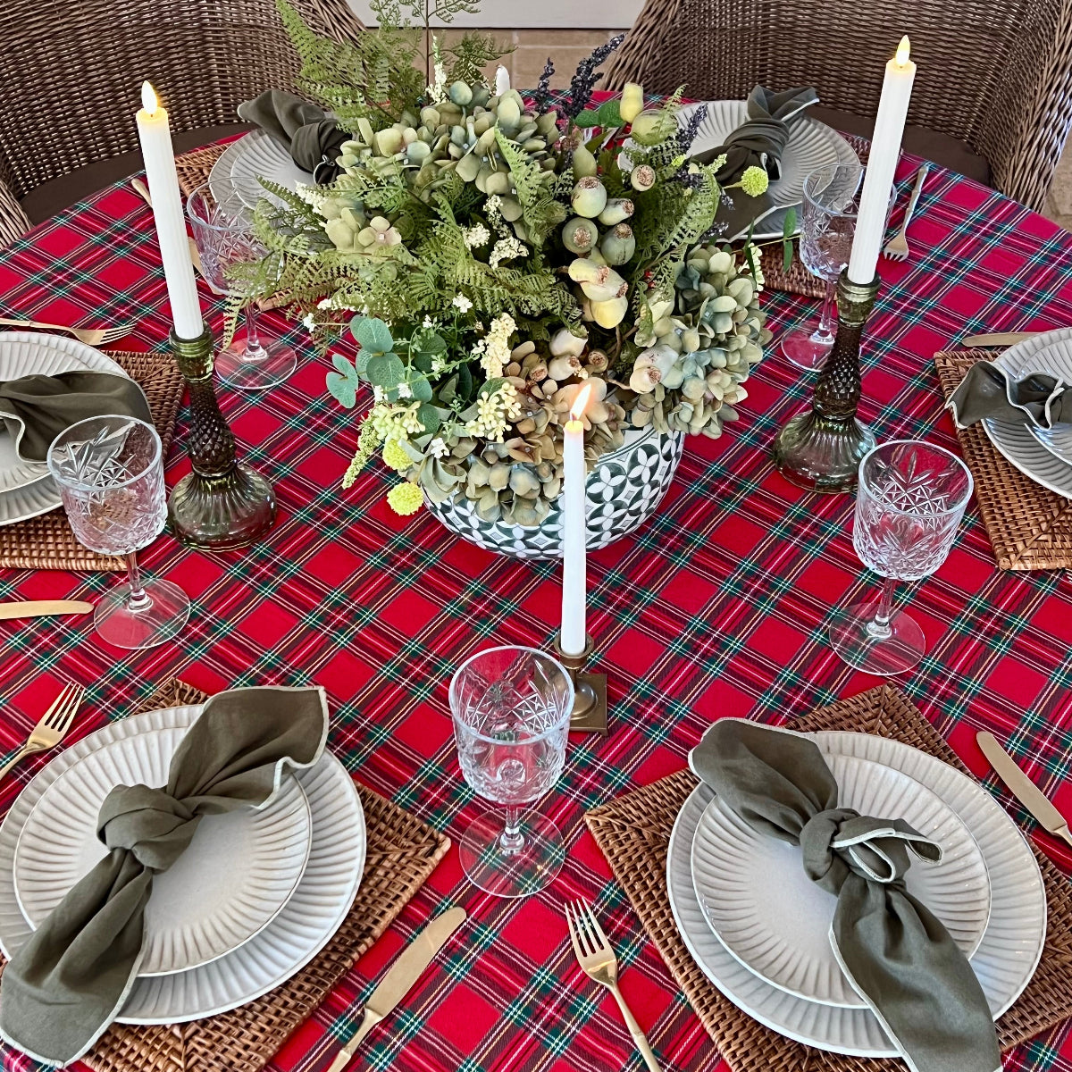 A festive dining table set with a red tartan plaid tablecloth, white plates, olive green napkins, glassware, and a large green floral centerpiece in a blue patterned vase, surrounded by wicker chairs.