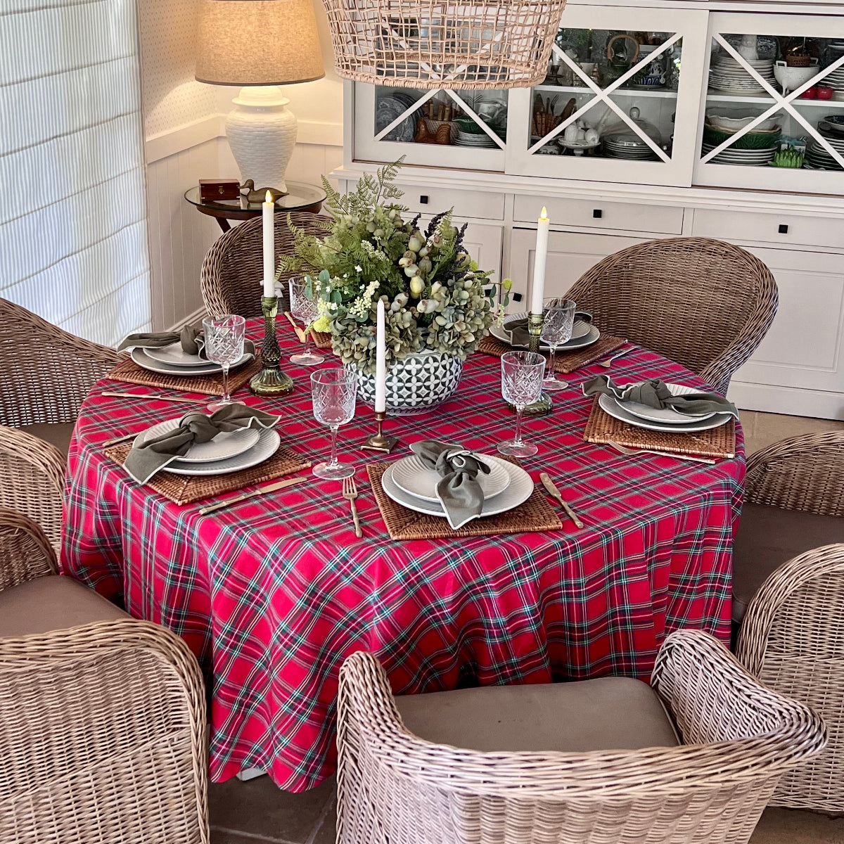 Round red tartan plaid tablecloth set for a cosy holiday dinner with white dinnerware, wine glasses and candlesticks