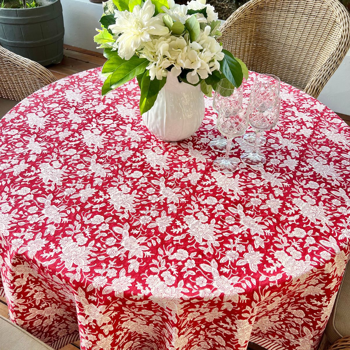 A red and white hand block printed floral patterned round tablecloth on a table, with a vase of flowers and glasses set on top.