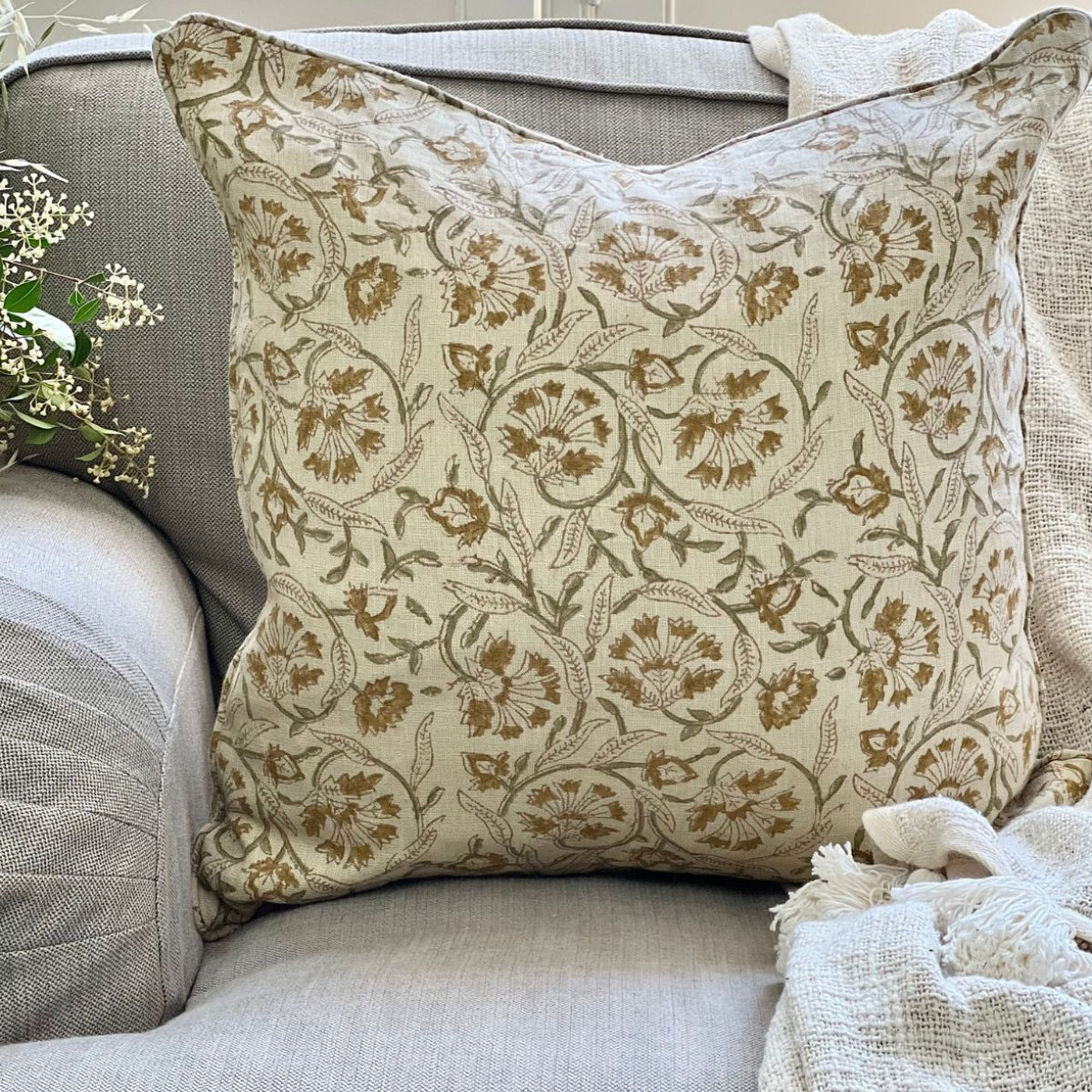 
A 50 x 50 cm Sunlit Vine floral hand block printed pure linen cushion cover with pipe edge finish, styled upfront on a sofa with a coffee and floral vase.