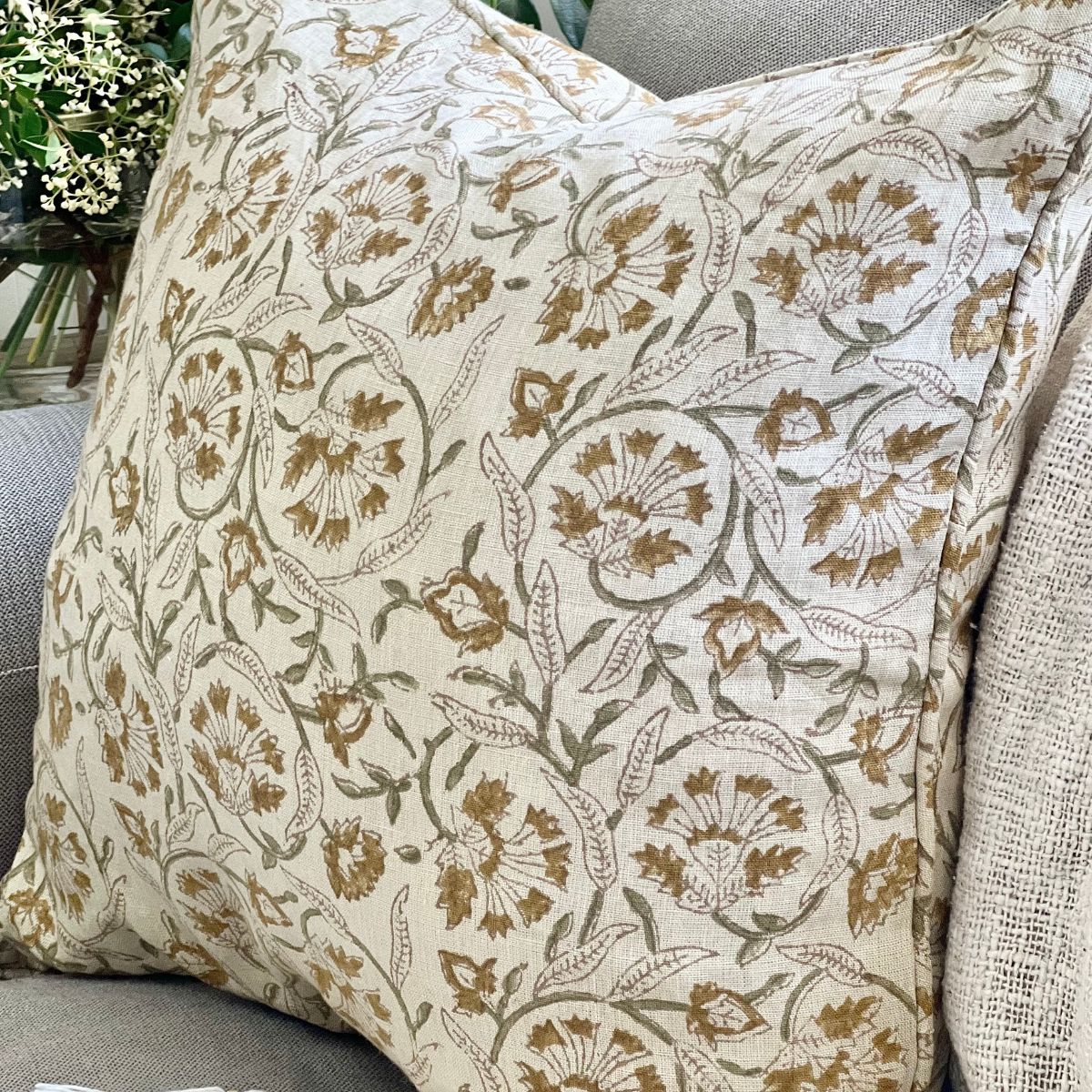 
A Sunlit Vine hand block printed pure linen cushion cover with zip closure and pipe edge finish, angled on a sofa showing the floral pattern with sage green vines across the linen fabric.