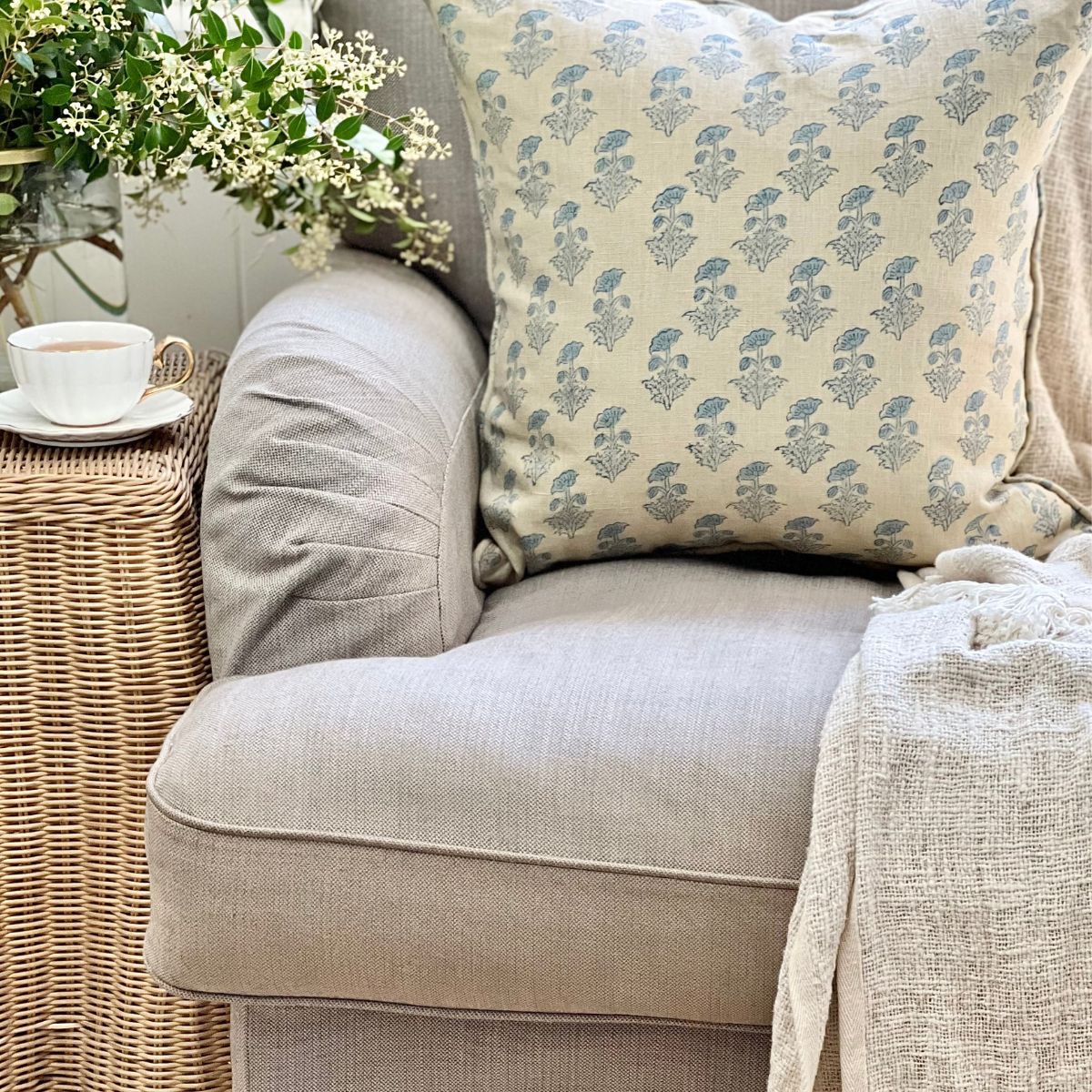 
A 50 x 50 cm Tranquil Blue floral hand block printed pure linen cushion cover with pipe edge finish, styled upfront on a sofa with a coffee and floral vase.