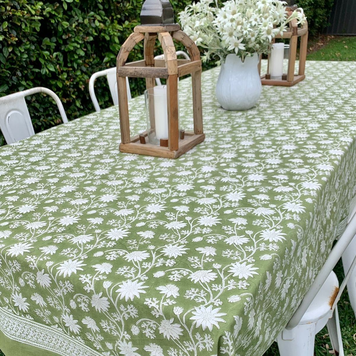 A green square tablecloth with a white flannel floral hand block printed pattern displayed on a table outdoors, with a wooden lantern and white chairs around it.