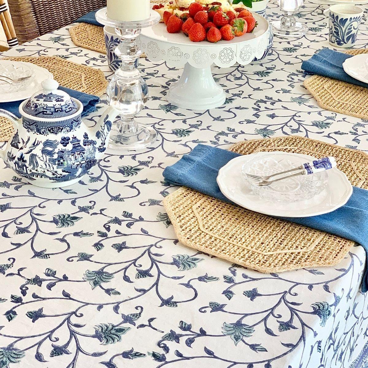 Side view of a round blue and green Ivy hand block printed cotton tablecloth on a circular dining table, with white plates and a lush floral vase centrepiece of garden blooms, the ivy print draping elegantly to the table edge.