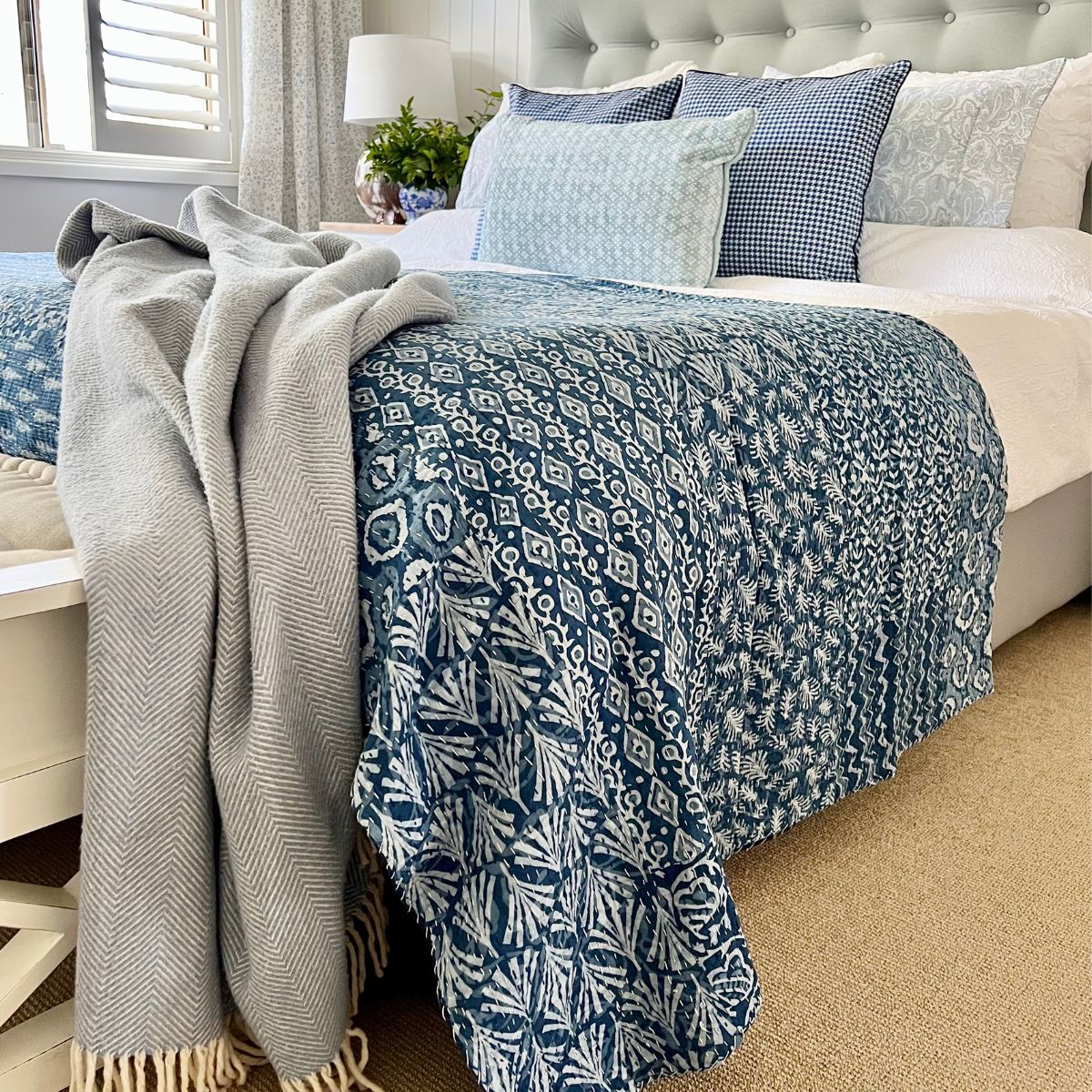 A blue and white patchwork kantha quilt spread out at the foot of a queen bed with various pillows on the bed, set in a bedroom with a nightstand, lamp, and plant decor.