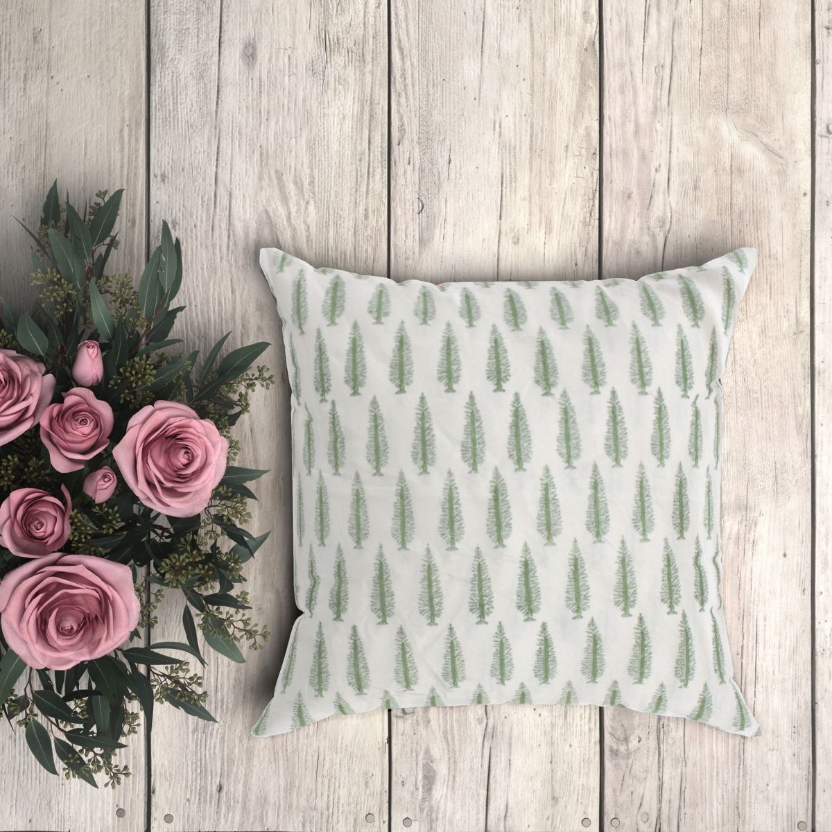 A green conifer tree cushion cover with a hand block printed pattern, displayed on a wooden surface next to a bouquet of pink roses.