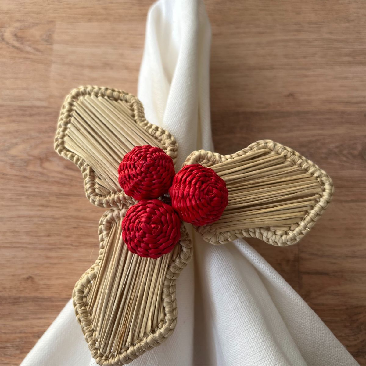 A red napkin ring with a holly berry design, consisting of red and beige accents, displayed on a white plate with a green patterned napkin.
