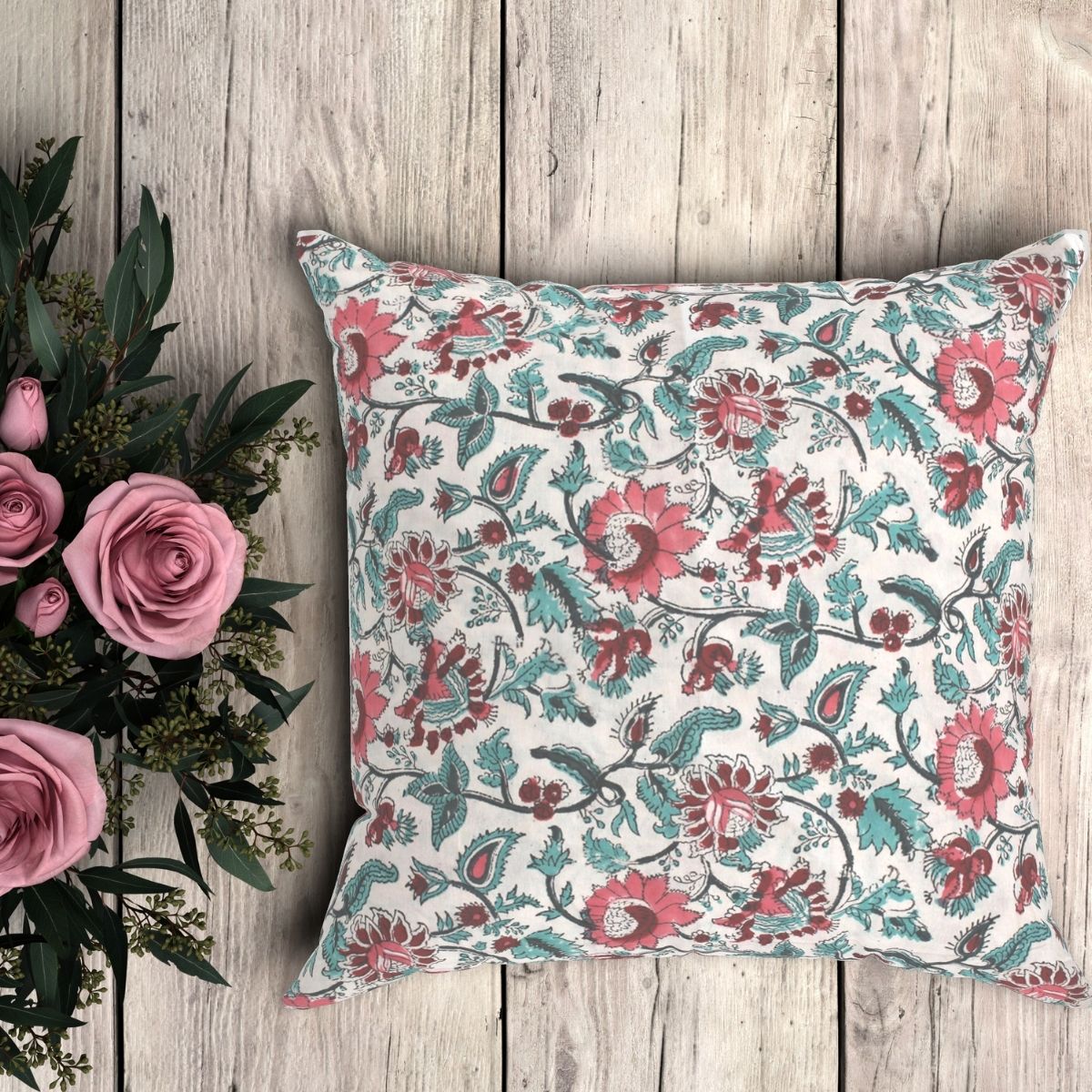 A pink blue hand block printed floral pattern cushion cover with a hand block printed pattern, displayed on a wooden surface next to a bouquet of pink roses.