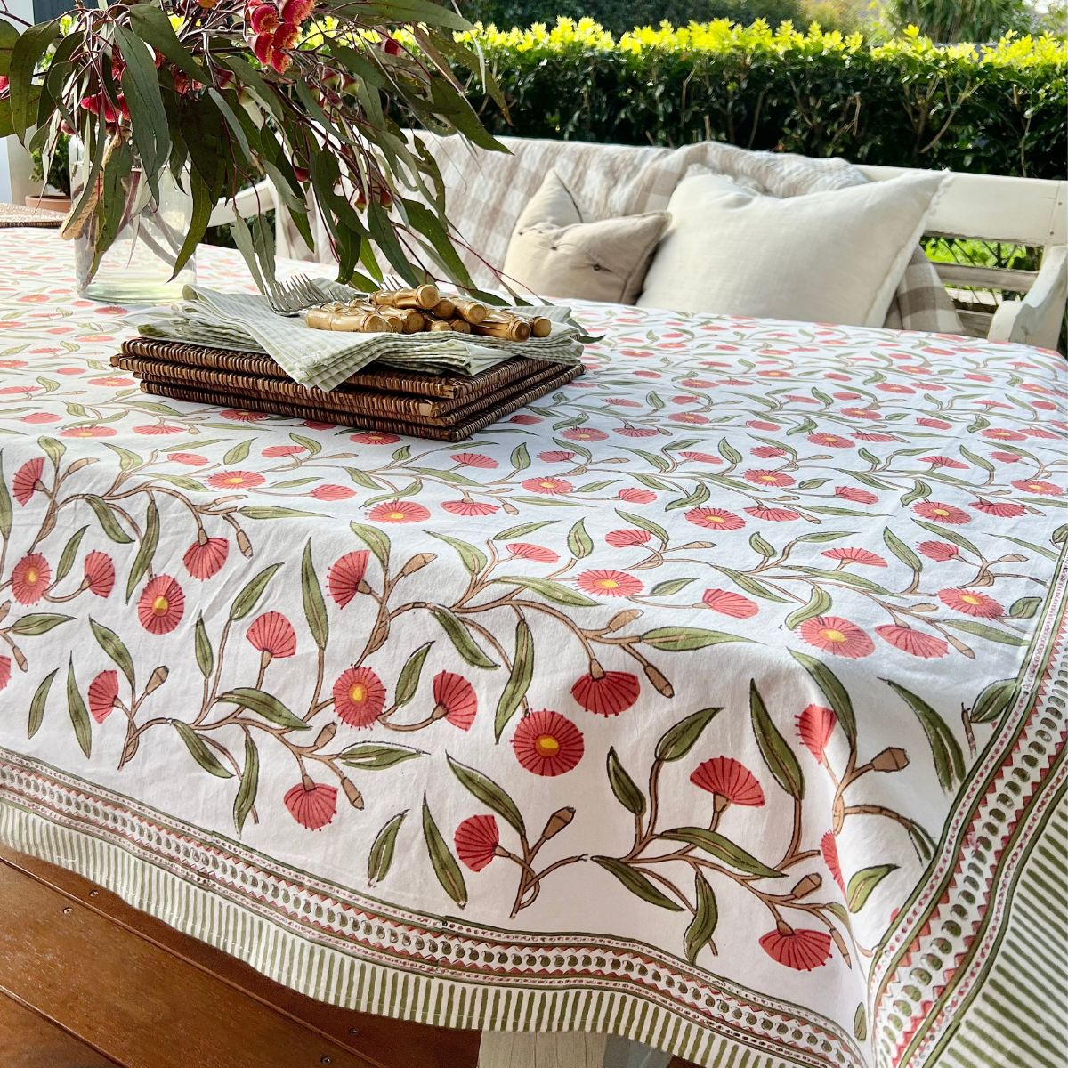 A square hand block printed tablecloth with a red flowering gum design displayed on a table outdoors.