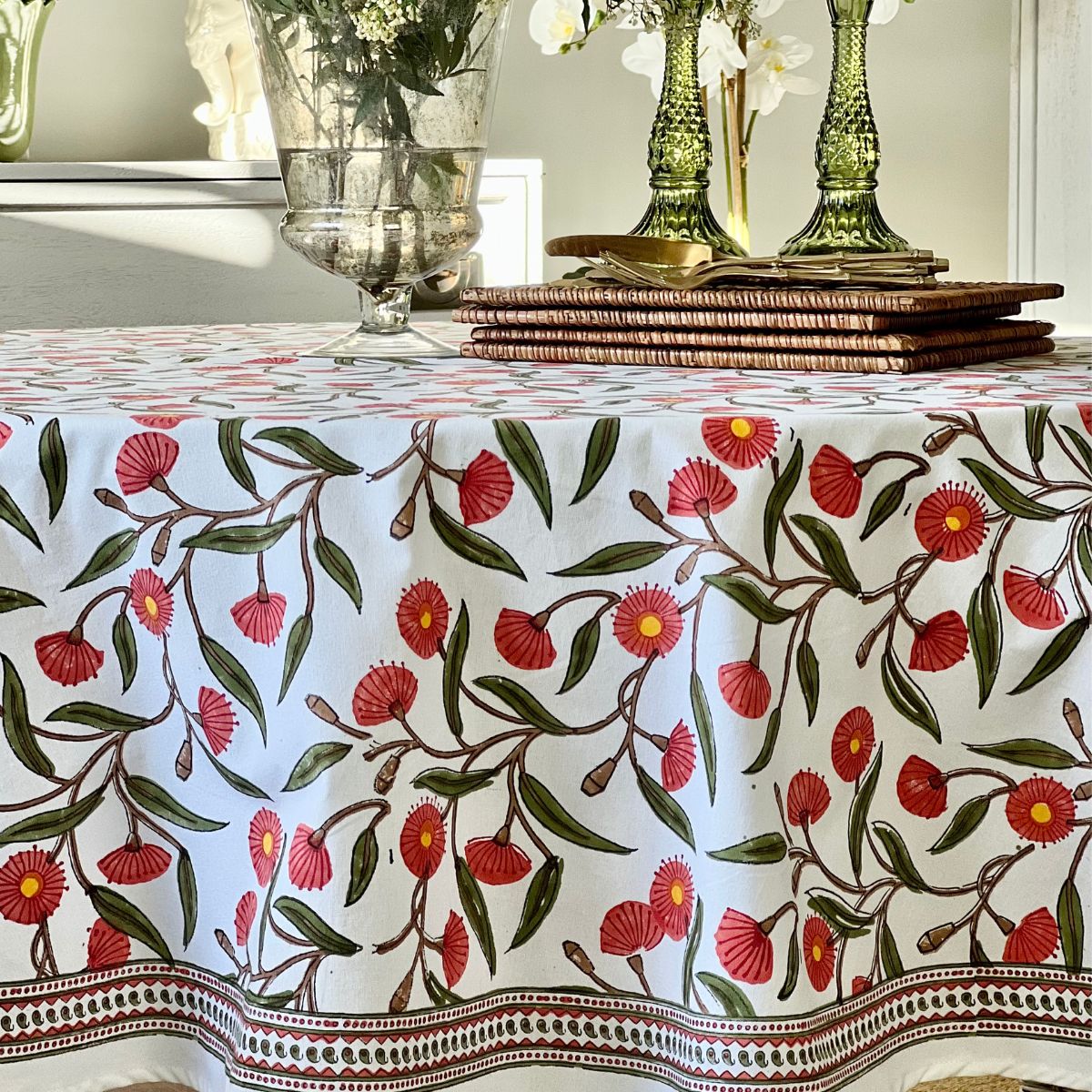 A round tablecloth with a red green flowering gum hand block printed design displayed on a table, paired with glassware and a white wicker chair in the setting.