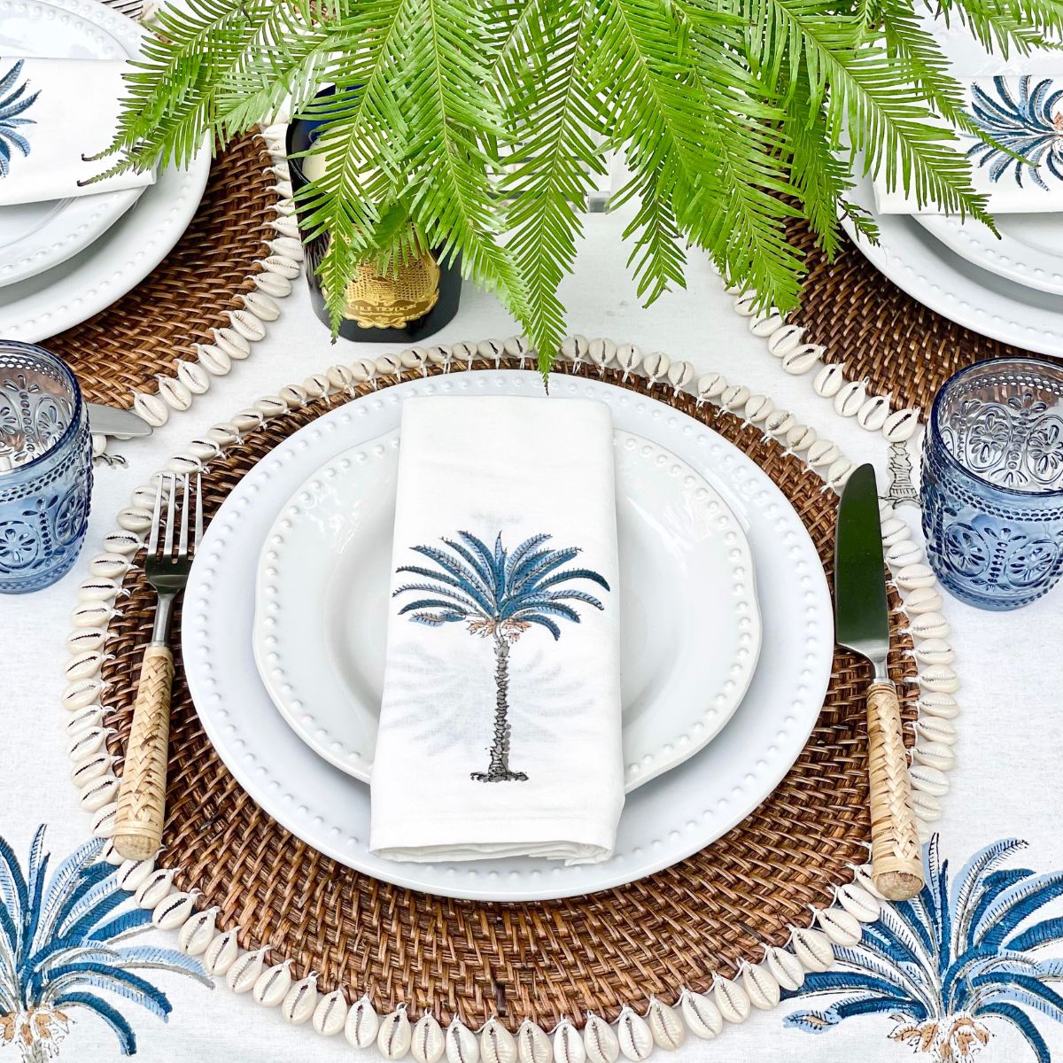 A round hand block printed tablecloth with a blue palm tree pattern displayed on a table outdoors, surrounded by plants.