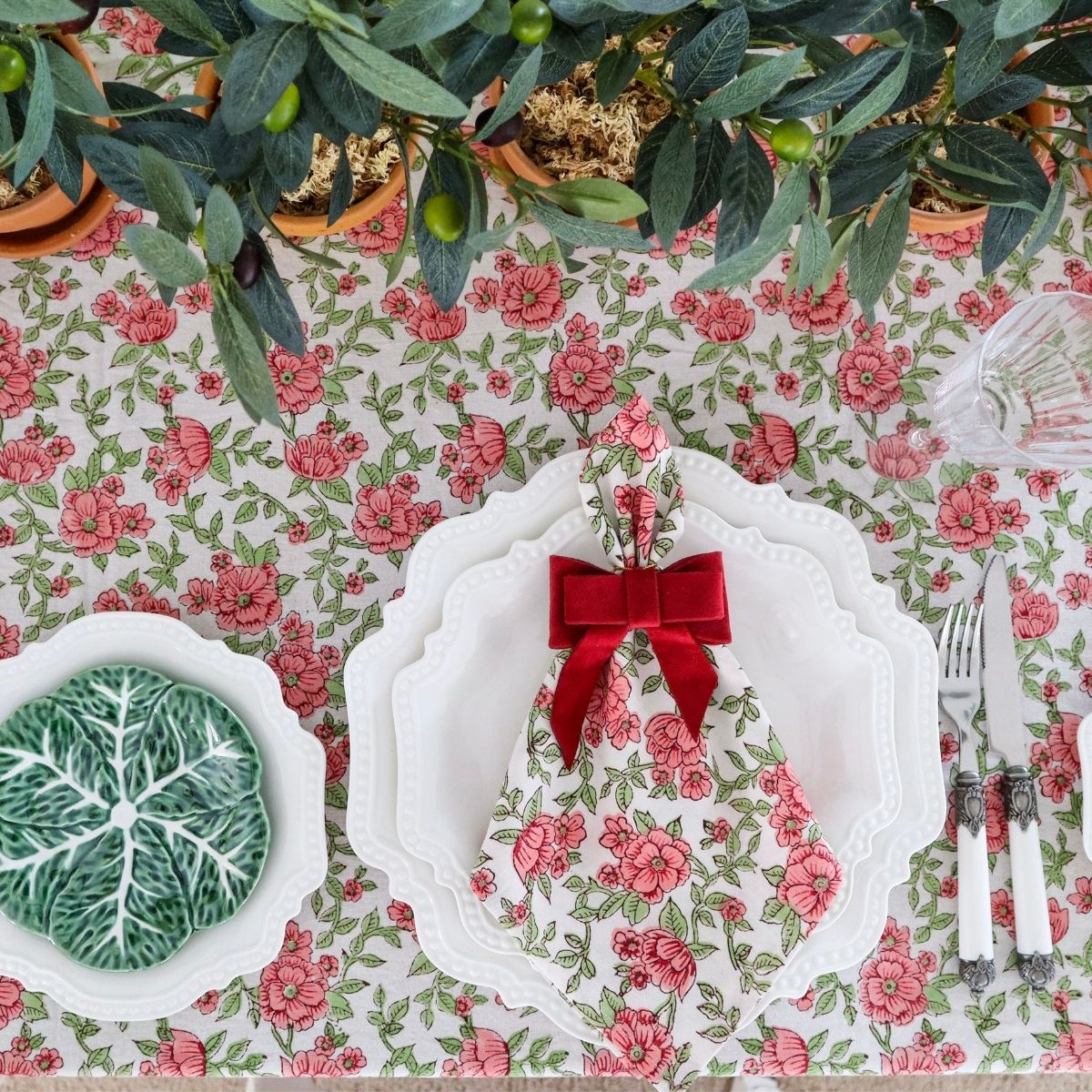 A set of white cotton table napkins with a red pink green hand block printed floral print and a red bow, displayed on a dining table with cutlery and a green plant in the background.