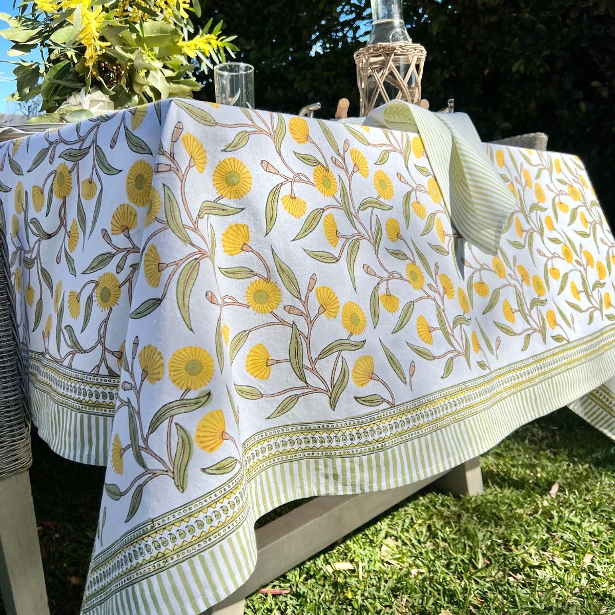 180 cm Yellow Flowering Gums square tablecloth styled with stacked dishes and cutlery for a relaxed outdoor garden picnic lunch.